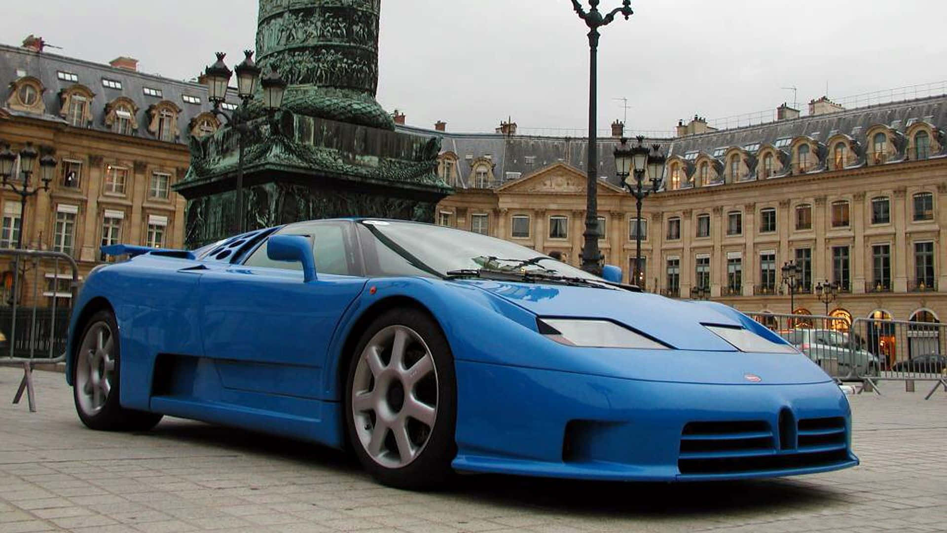 Bugatti Eb110 桌布 壁紙 — 一輛充滿活力的藍色布加迪EB110超級跑車停泊在鵝卵石廣場上，前方是宏偉的歷史紀念碑和古典建築，天空多雲。