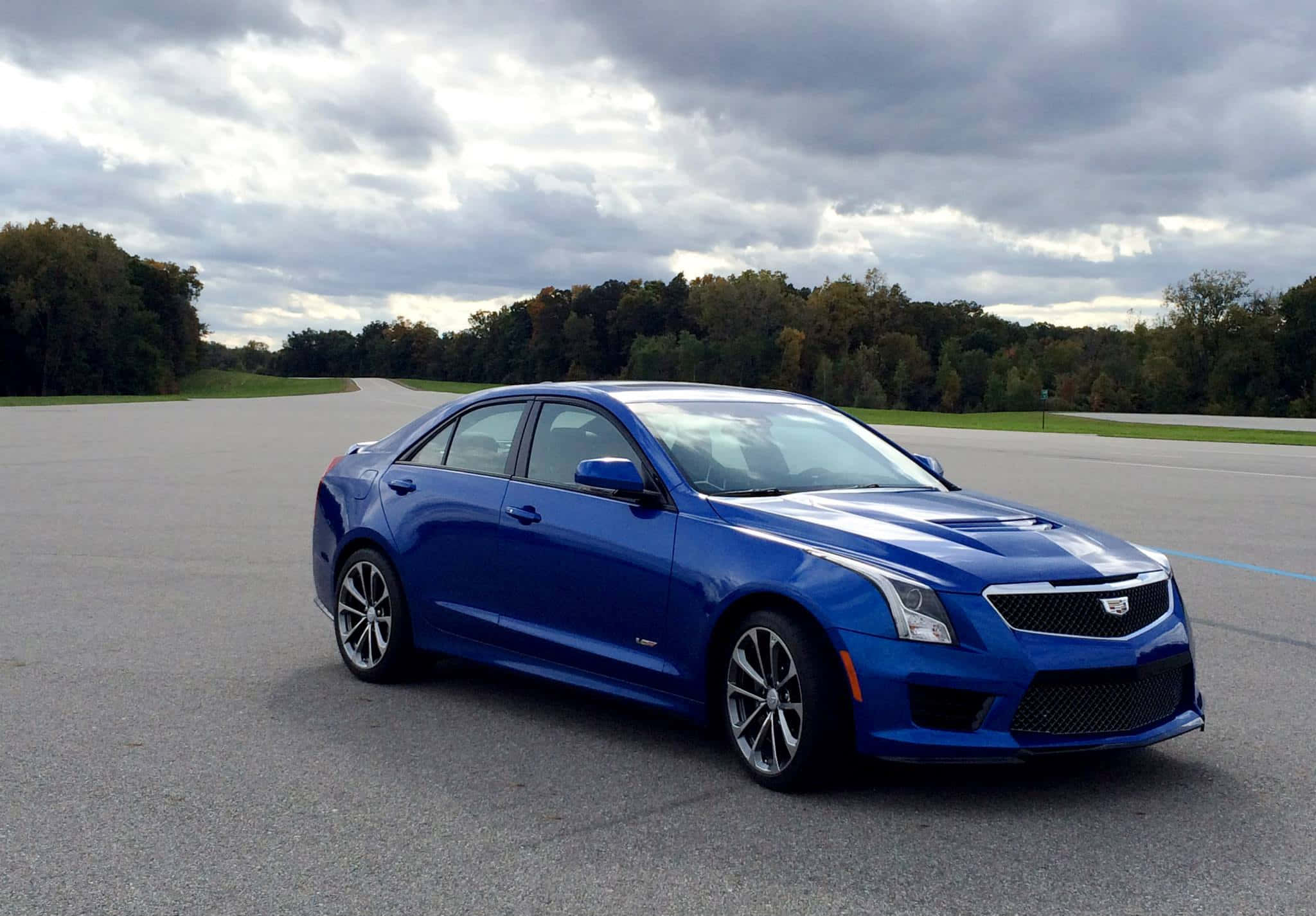 Cadillac Ats 桌布 壁紙 — 一輛充滿活力的藍色Cadillac ATS-V轎車在陰天之下，於一條兩旁種滿秋樹的空蕩瀝青路上閃閃發光。
