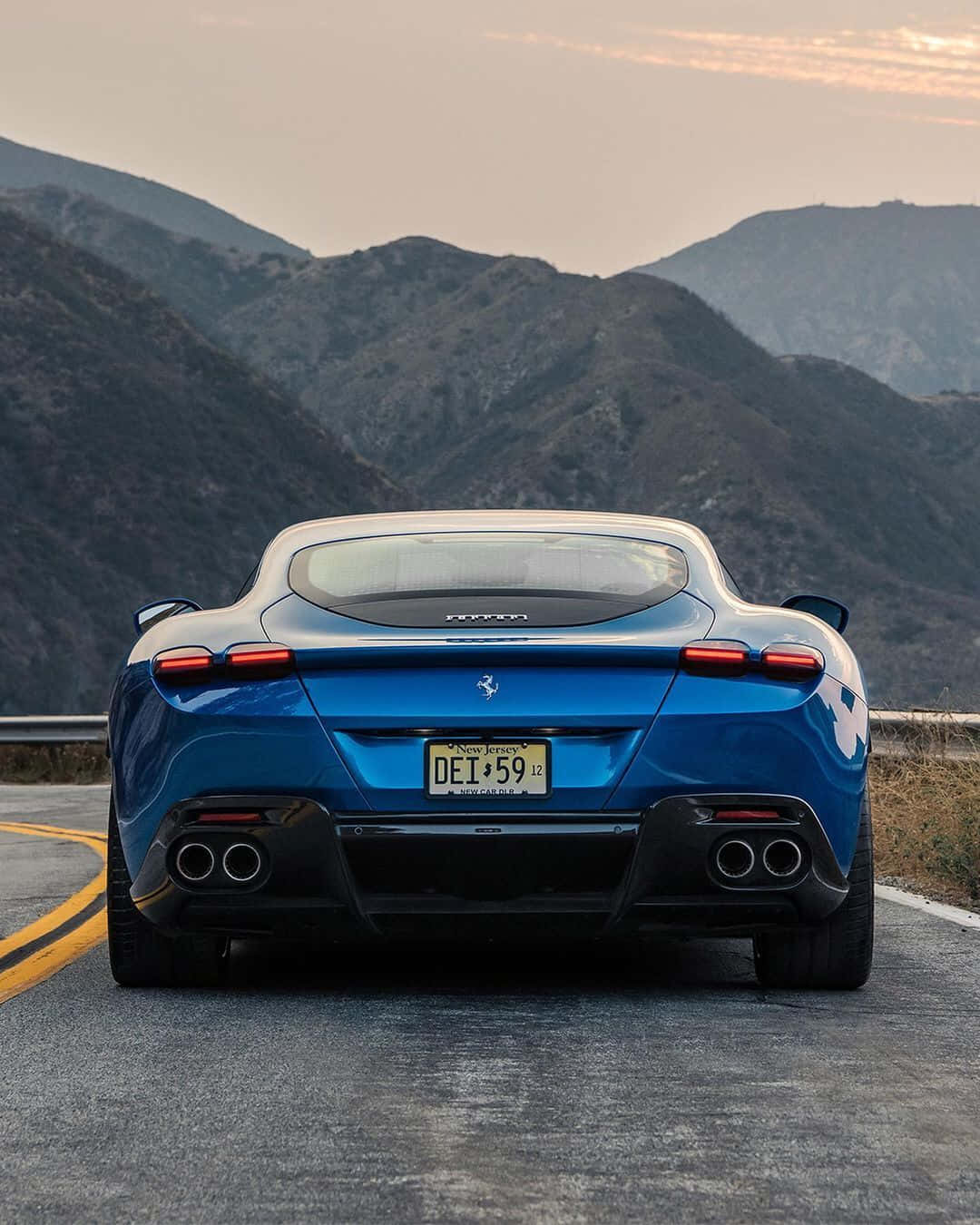 Ferrari Roma 桌布 壁紙 — 一輛充滿活力的藍色法拉利在日落時分沿著蜿蜒的山路行駛，背景是崎嶇的山峰。