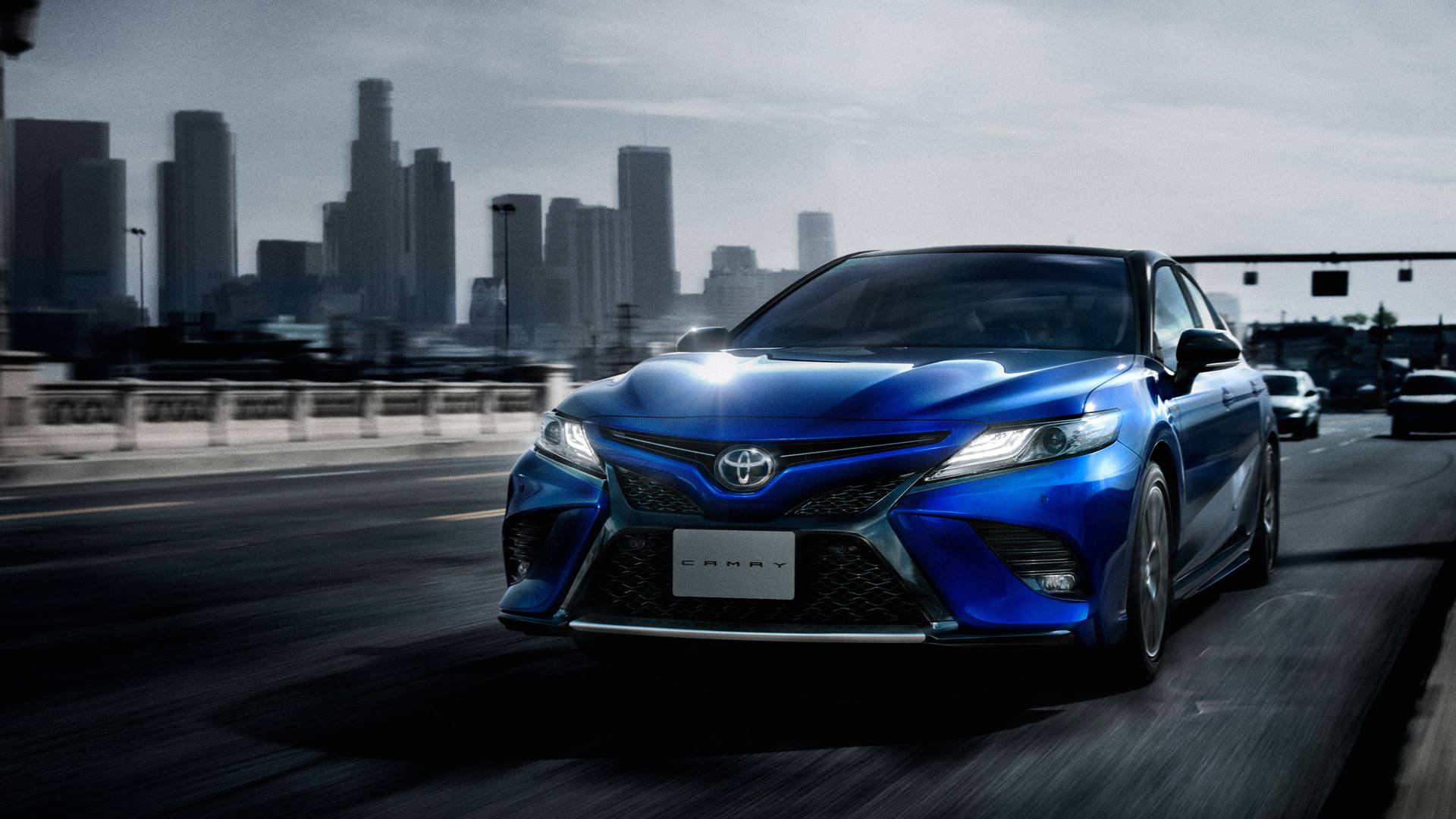 藍色豐田Camry在城市高速公路上疾馳，背景有摩天大樓。 — Toyota Supra 壁紙