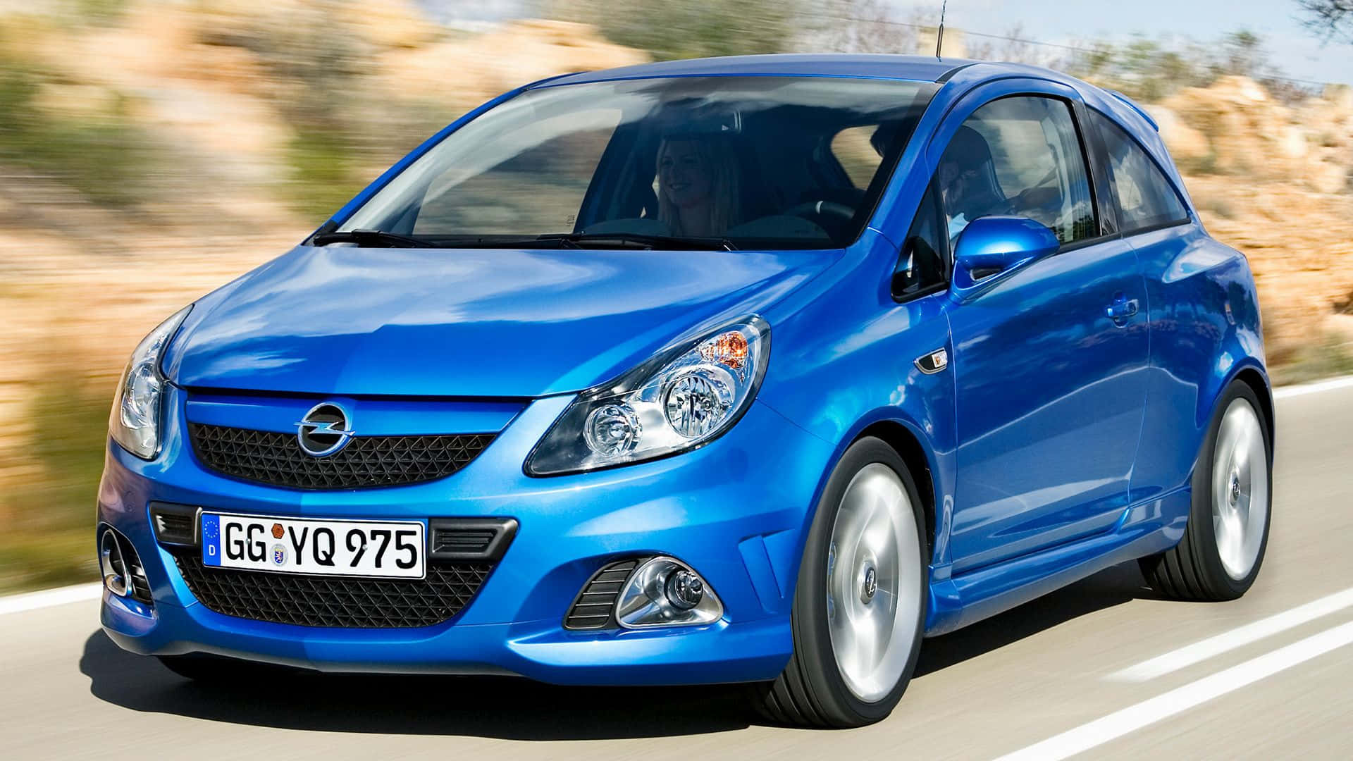 Opel Corsa 桌布 壁紙 — 一輛充滿活力的藍色Opel Corsa在陽光明媚的道路上行駛，背景是模糊的岩石地形。