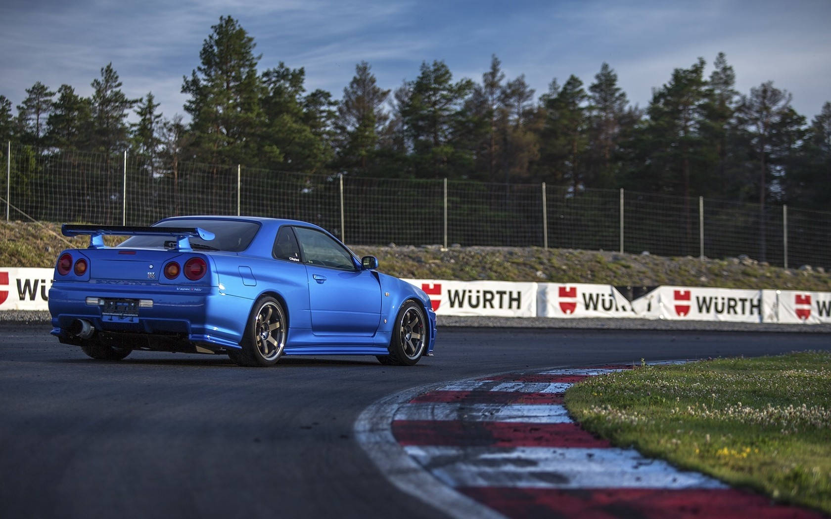 藍色日產Skyline R34在賽道上飄移，賽道有紅白路緣。 — 天際線 壁紙