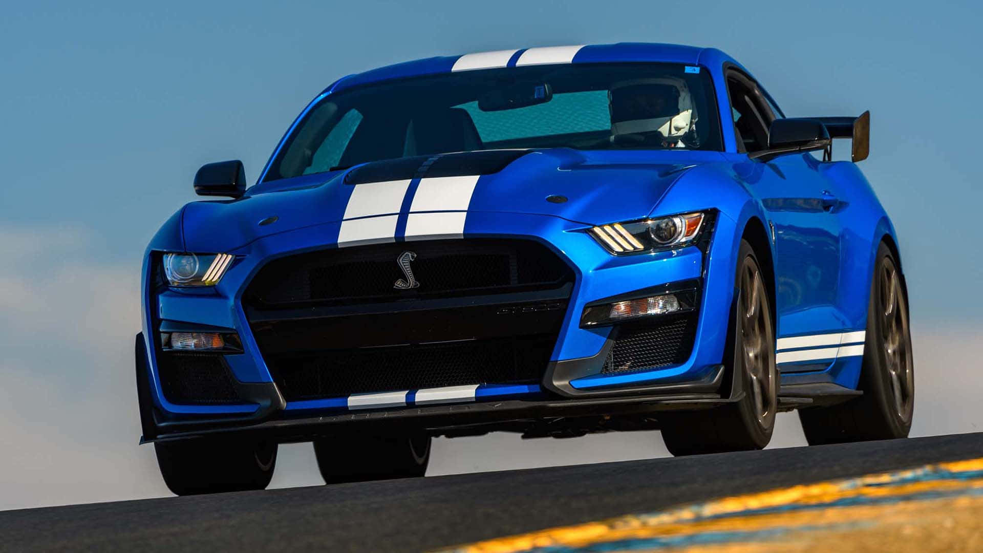  壁紙 — 一輛充滿活力的藍色Shelby GT500跑車，帶有白色賽車條紋，在晴朗的天空下沿著賽道飛馳。