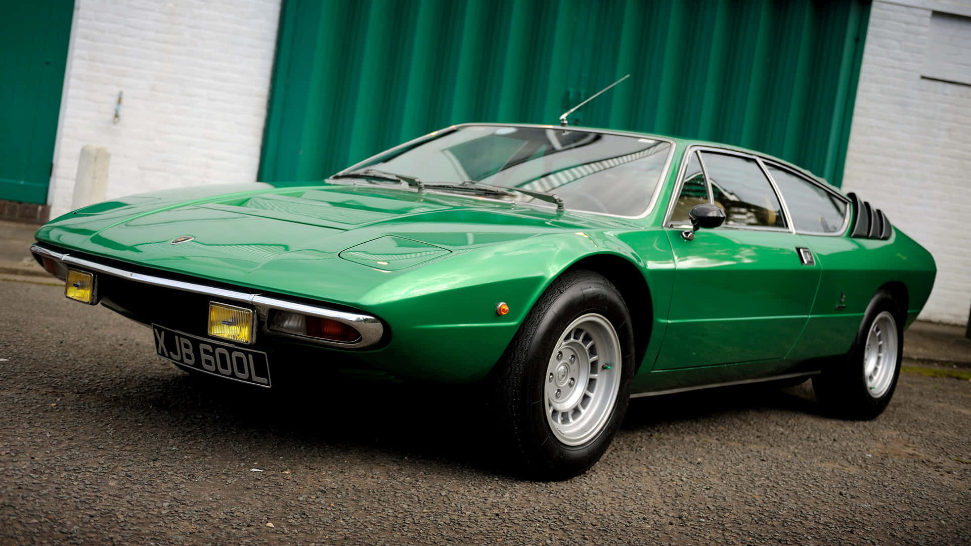 Lamborghini Urraco 桌布 壁紙 — 一輛充滿活力的綠色經典跑車停在白色磚牆和綠色波紋門前的瀝青路面上。