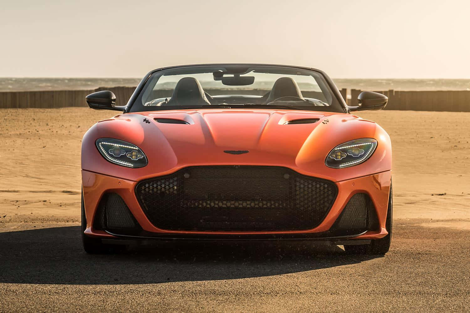 Aston Martin Dbs Superleggera 桌布 壁紙 — 一輛充滿活力的橙色 Aston Martin DBS Superleggera 敞篷車停放在陽光明媚的沙漠道路上，有木柵欄和遙遠的地平線。