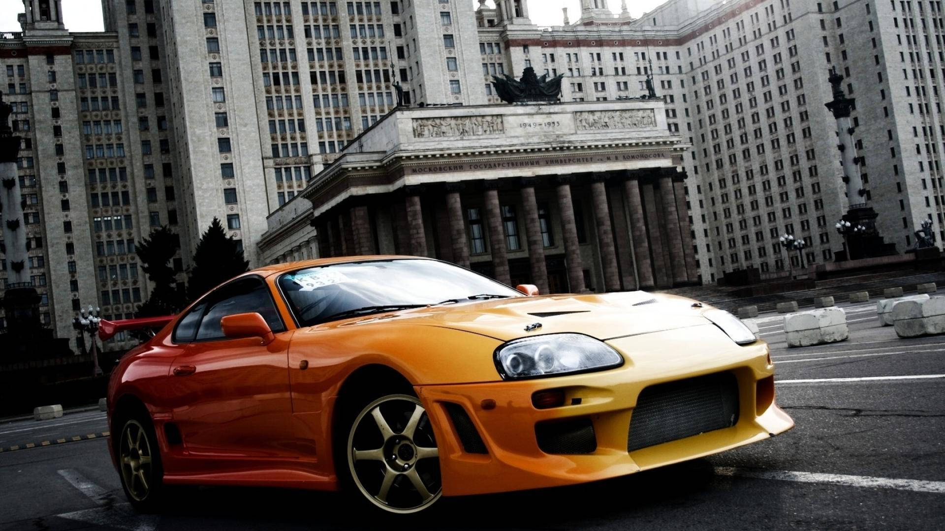 日落時分，橙色跑車停泊在一座宏偉的城市建築前。 — Toyota Supra 壁紙