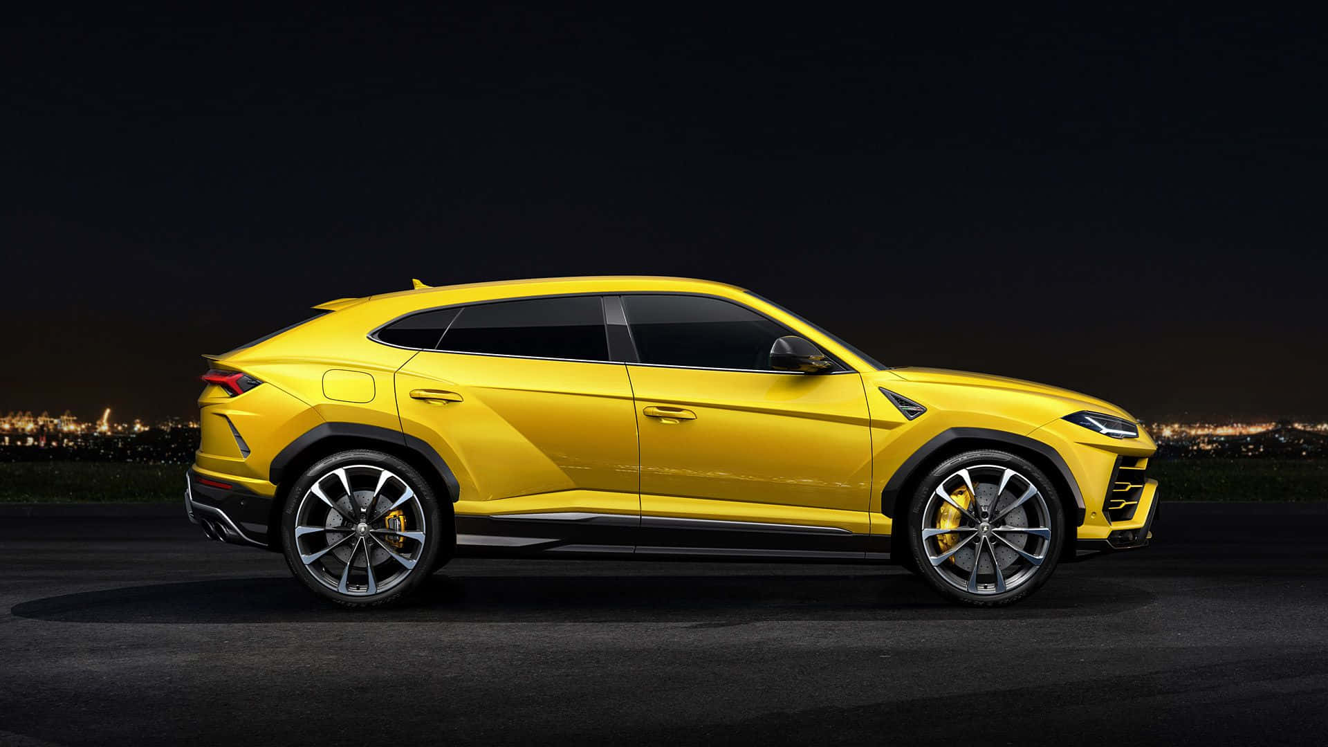 Lamborghini Urus 桌布 壁紙 — 一輛充滿活力的黃色藍寶堅尼 Urus SUV 在夜燈下閃閃發光，遠處的城市天際線閃爍著光芒。