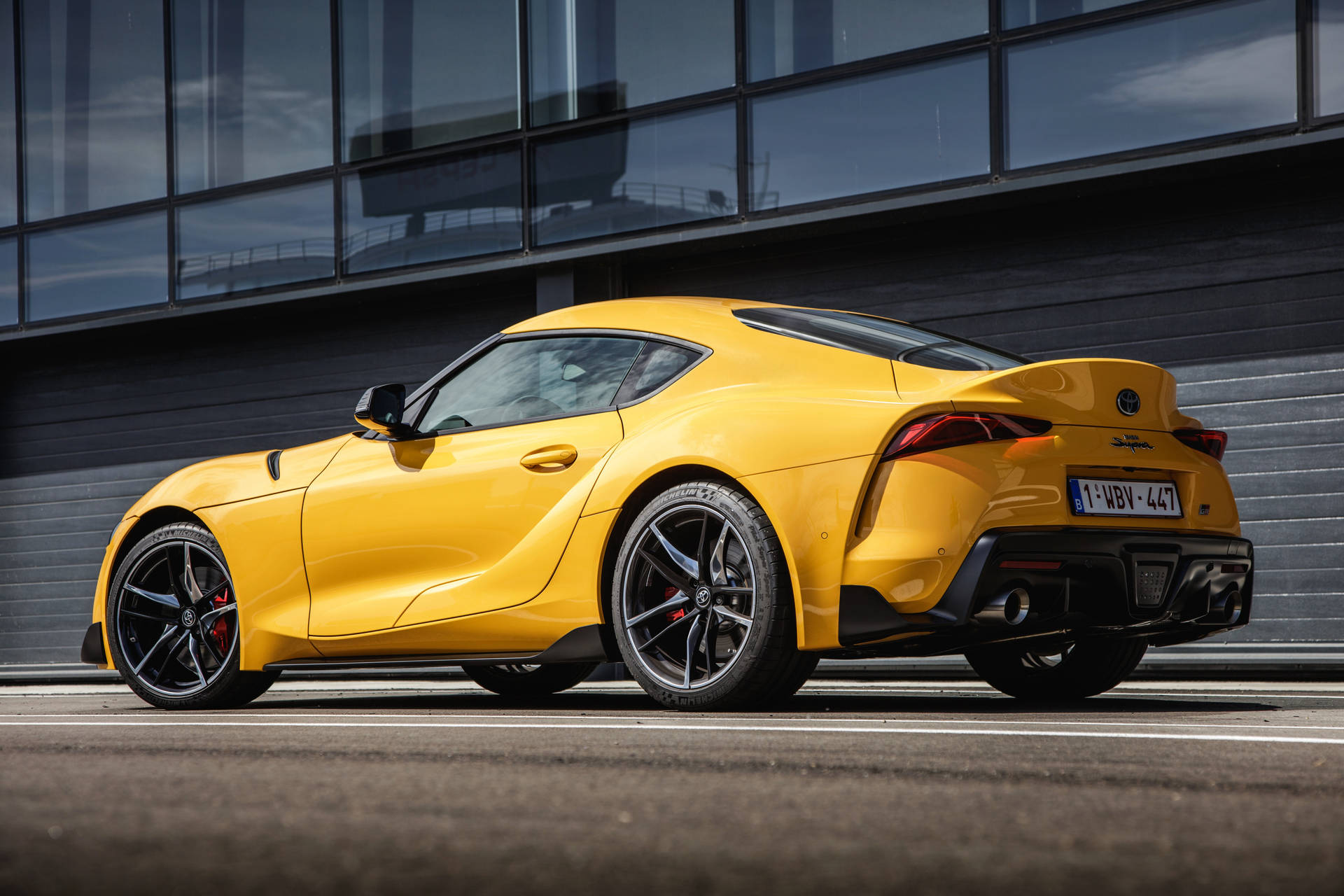 黃色跑車停在一棟現代玻璃建築前。 — Toyota Supra 壁紙