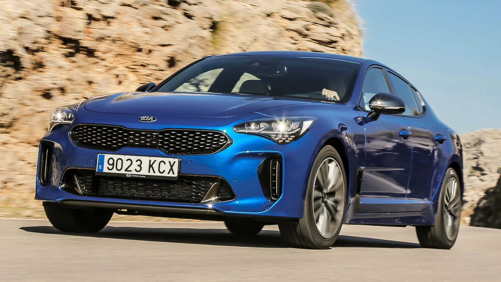 Kia Stinger 桌布 壁紙 — 一輛充滿活力的藍色Kia Stinger跑車在陽光明媚的山路上飛馳，背景是岩石峭壁。