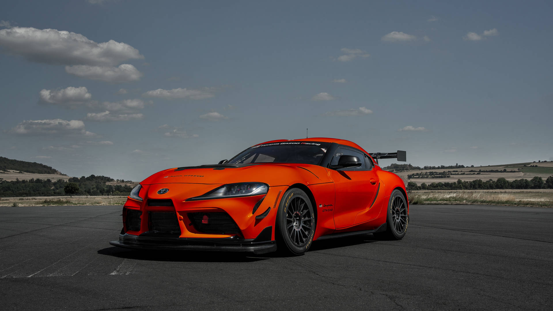 橙色賽車在瀝青賽道上，背景有丘陵和日落天空 — Toyota Supra 壁紙