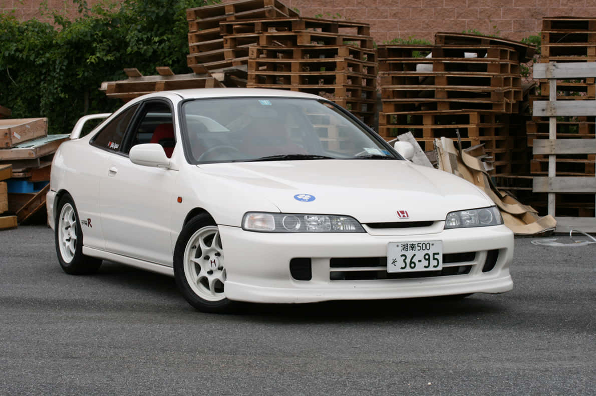 Honda Integra 桌布 壁紙 — 一輛純白色本田Integra Type R雙門跑車停在柏油路上，背景有堆疊的木棧板和綠植。
