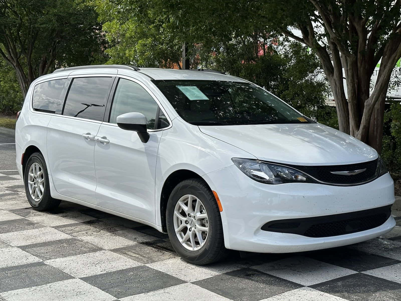 Chrysler Voyager 桌布 壁紙 — 一輛純白色的 Chrysler Pacifica 小型貨車停放在黑白棋盤格路面上，上方有綠葉成蔭的樹木。