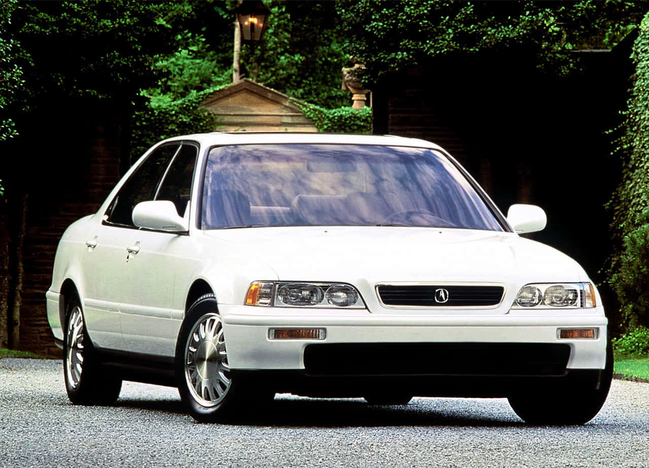 Acura Legend 桌布 壁紙 — 一輛純白色的Acura轎車停放在碎石車道上，周圍環繞著茂密的綠植和一座石屋。