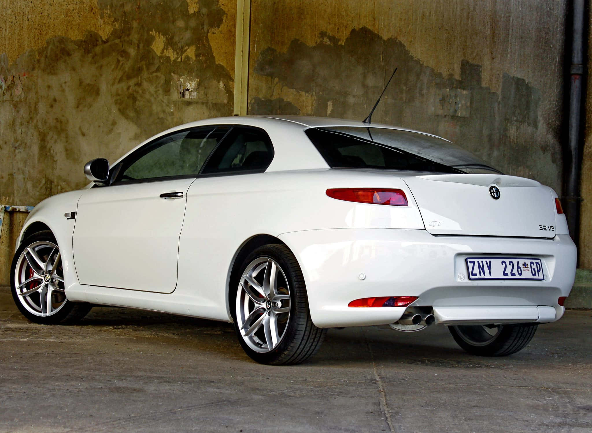 Alfa Romeo Gt 桌布 壁紙 — 一輛純白色的愛快羅密歐GT轎跑車停放在光線昏暗的車庫中，靠著風化的混凝土牆。