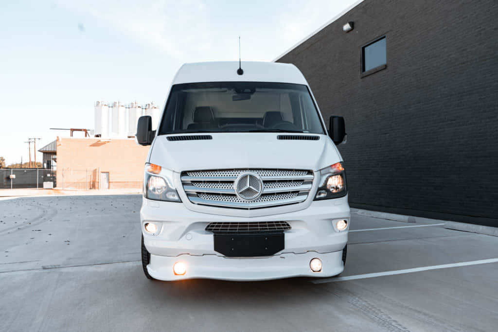 Mercedes Benz Sprinter 桌布 壁紙 — 一輛純白色的 Mercedes-Benz Sprinter 客貨車停放在混凝土停車場，旁邊是深色磚砌建築，天空晴朗。