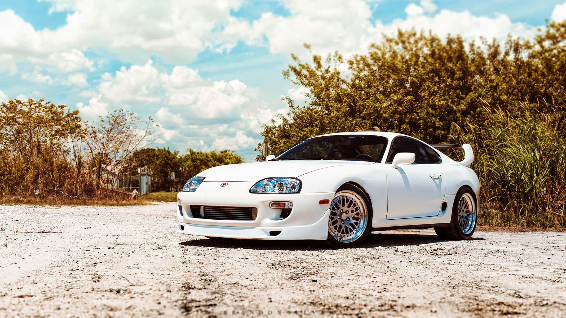白色豐田Supra跑車搭配藍色輪圈，停放在樹旁的碎石上。 — Toyota Supra 壁紙