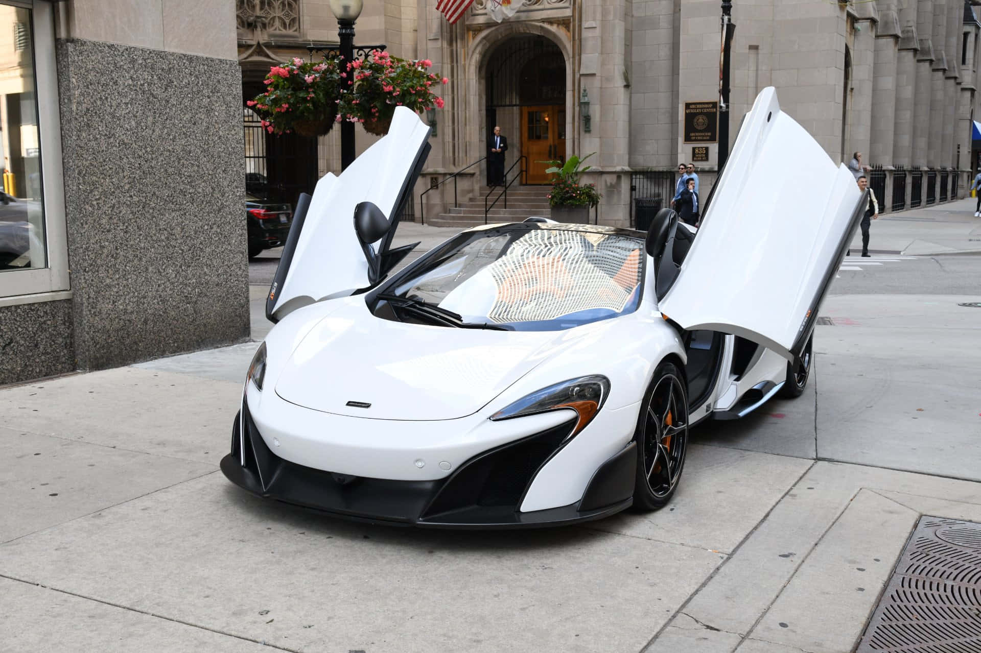 Mclaren 675lt Spider 桌布 壁紙 — 一輛純白色McLaren跑車，鷗翼車門打開，停放在靠近掛有花籃的石建築物的城市人行道上。
