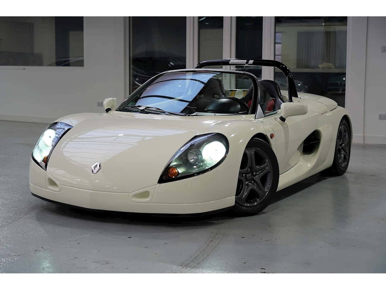 Renault Sport Spider 桌布 壁紙 — 一輛純淨的奶油色雷諾蜘蛛跑車，帶有黑色飾邊和發光的車頭燈，停在室內拋光混凝土地板上。