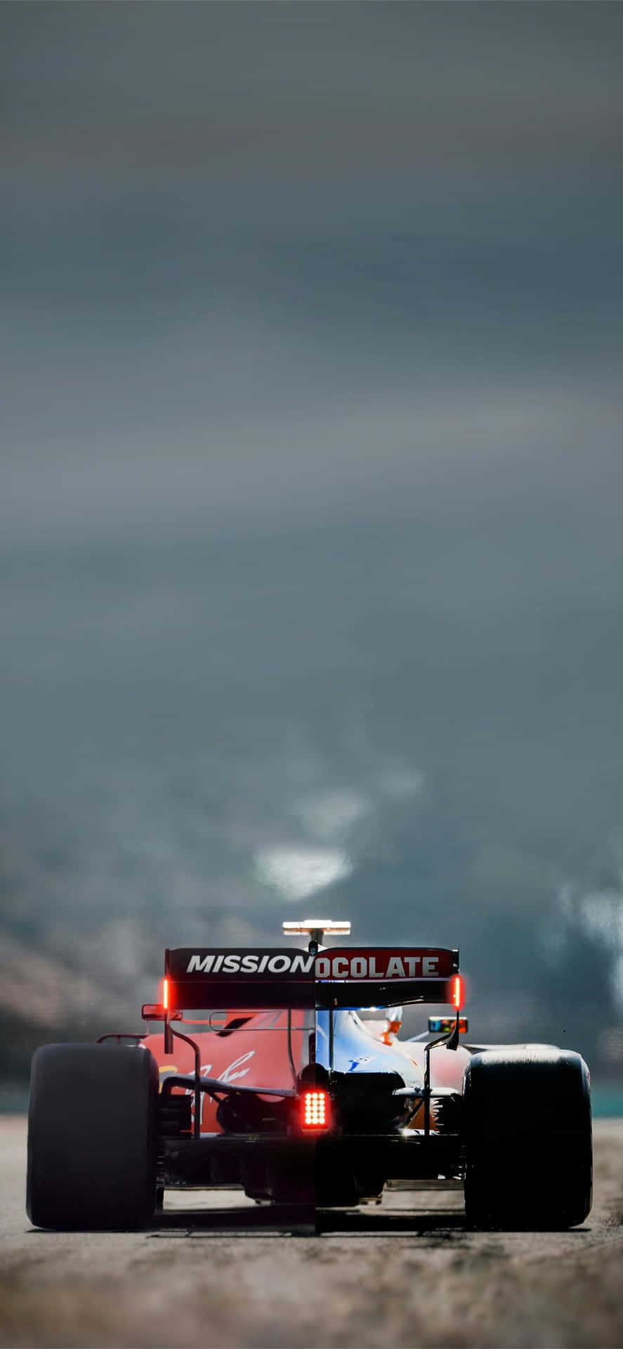  壁紙 — 一輛帶有「MISSION OCOLATE」品牌的紅色一級方程式賽車，在灰濛濛的天空下於賽道上奔馳。