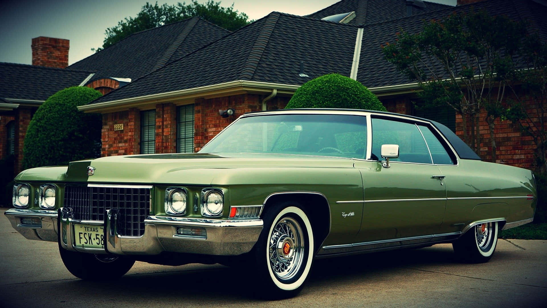 Cadillac Deville 桌布 壁紙 — 一輛復古橄欖綠色的凱迪拉克 Coupe de Ville，配備白色牆壁輪胎，停在一間磚房前，周圍有修剪整齊的樹籬。