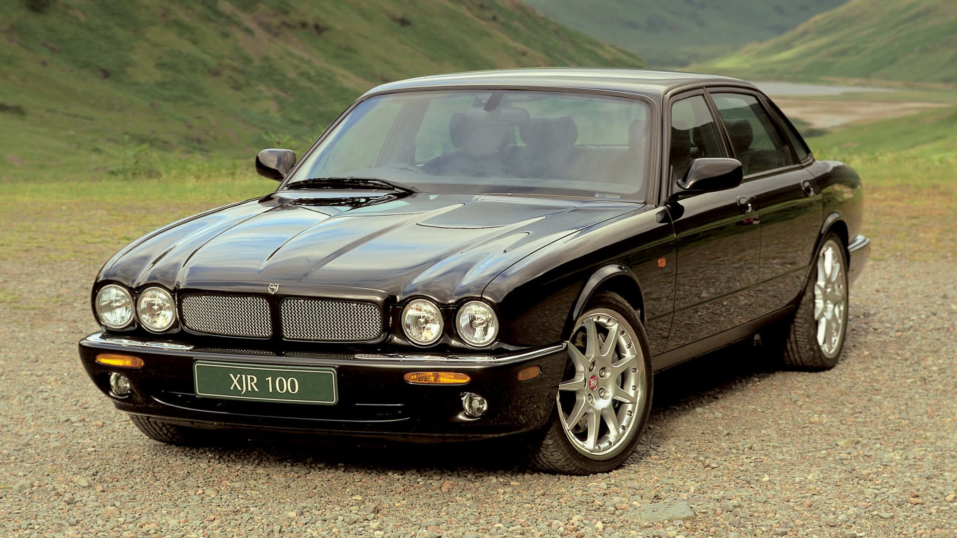 Jaguar Xjr 桌布 壁紙 — 一輛光滑的黑色積架XJR 100轎車在碎石路上閃閃發光，背景是起伏的綠色山丘。