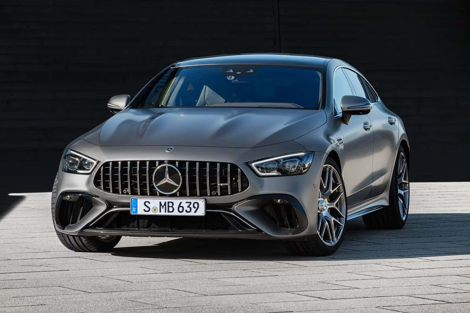 Mercedes Benz Amg Gt 桌布 壁紙 — 一輛光滑的啞光灰色梅賽德斯-AMG GT 63 S 轎跑車停在鋪砌路面上，背景是深色木牆，在明亮的日光下。