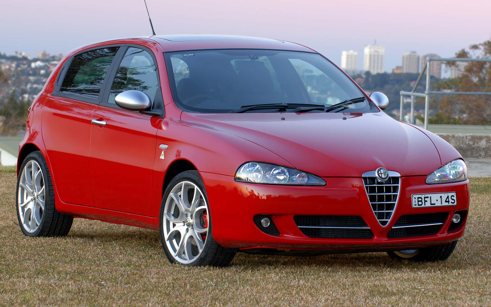 Alfa Romeo 147 桌布 壁紙 — 一輛光亮的紅色愛快羅密歐147跑車停在草地上，黃昏時分可見城市天際線。