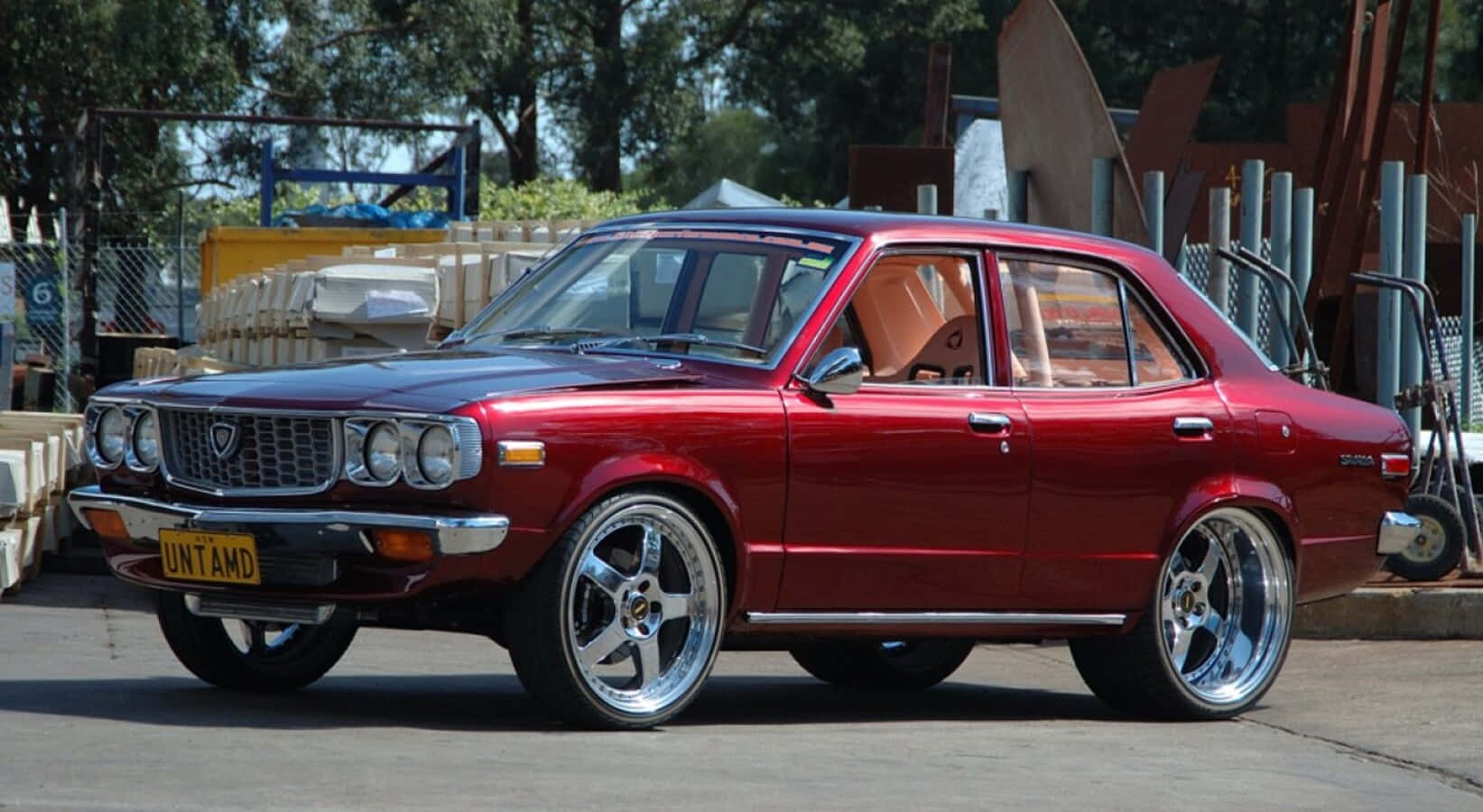 Mazda Rx-3 桌布 壁紙 — 一輛光亮的紅色經典轎車配備定制鍍鉻輪圈，停在混凝土上，靠近工業材料，在明亮的日光下。
