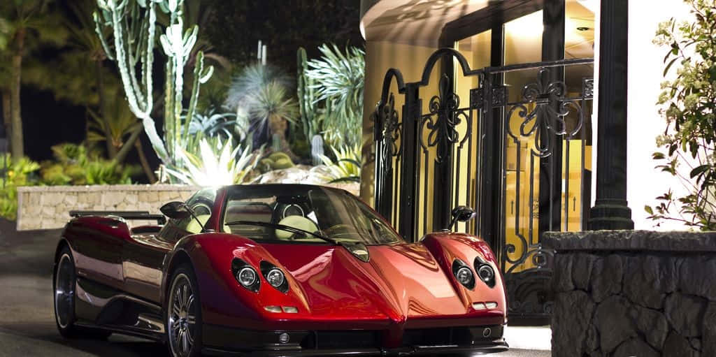 Pagani Zonda C12 Roadster 桌布 壁紙 — 一輛光亮的紅色帕加尼Zonda跑車在夜間停放在一個裝飾華麗的大門前，門旁有發光的仙人掌和棕櫚樹。