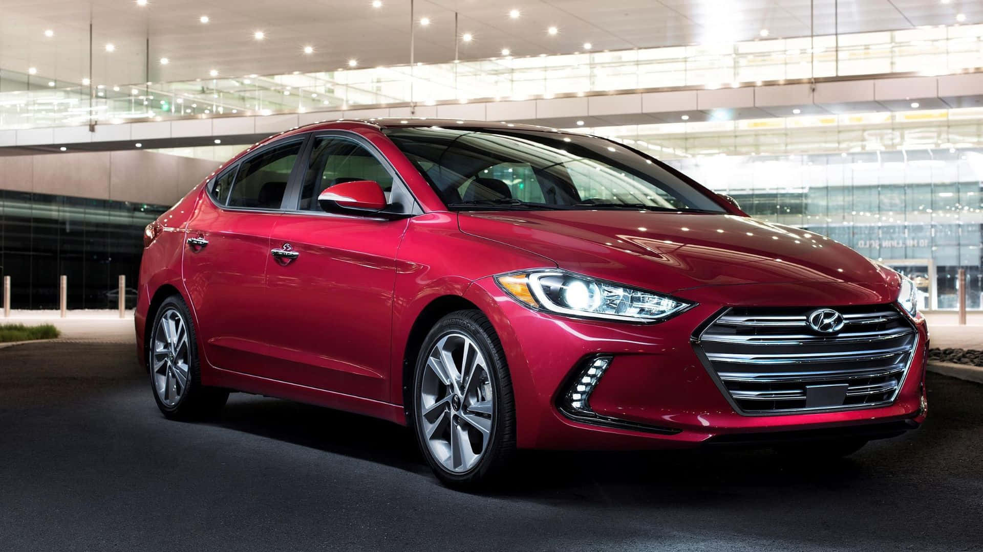 Hyundai Elantra 桌布 壁紙 — 一輛光亮的紅色現代轎車在現代玻璃建築停車區的明亮燈光下閃閃發光。