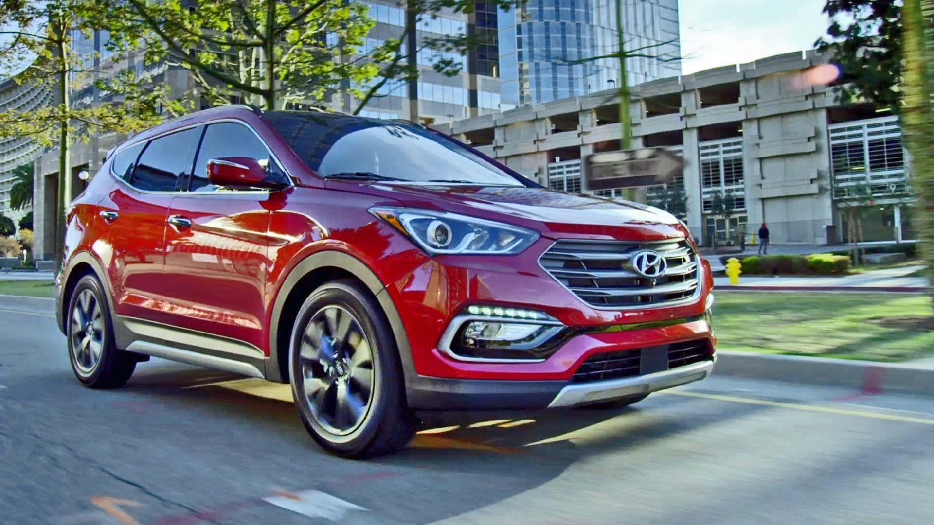Hyundai Santa Fe 桌布 壁紙 — 一輛光亮的紅色現代SUV在陽光明媚的城市街道上行駛，背景是現代建築和綠樹。