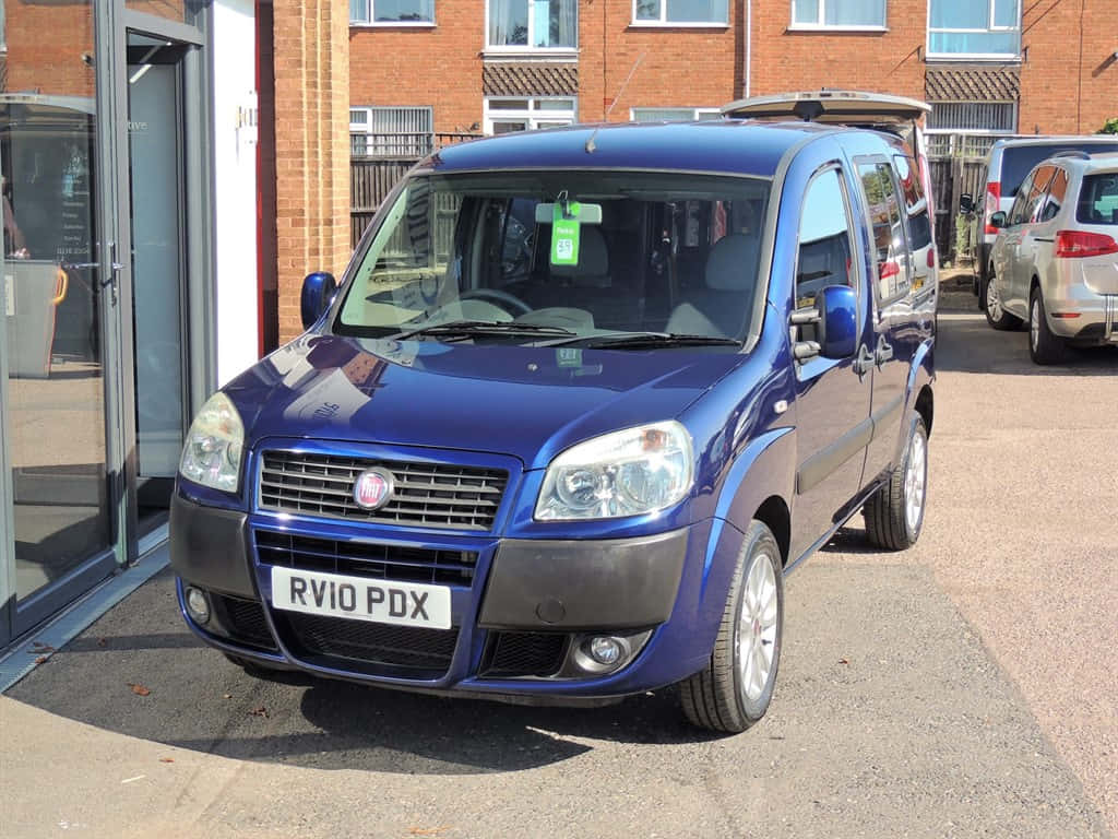  壁紙 — 一輛光亮的藍色Fiat Panda 4x4停放在鋪砌的停車場，旁邊是帶玻璃門的磚造建築。
