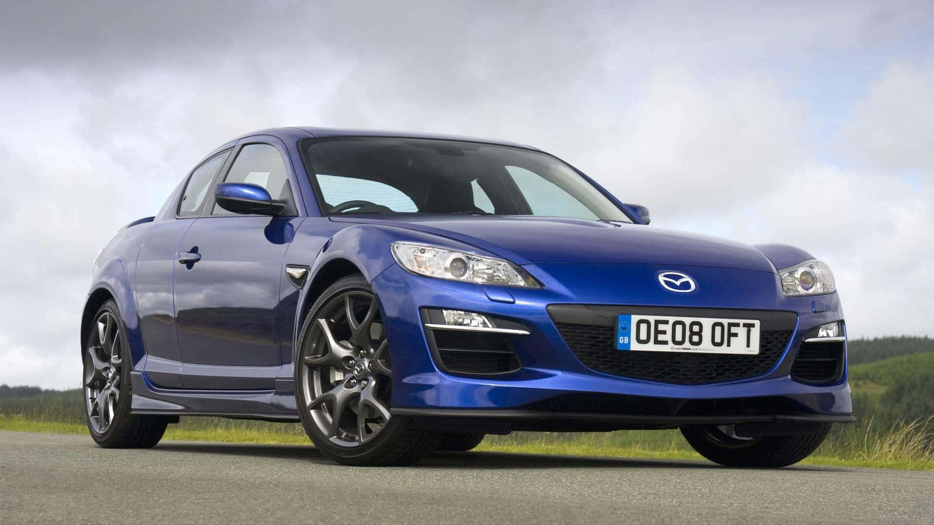 Mazda Rx-8 桌布 壁紙 — 一輛光亮的藍色馬自達RX-8跑車停泊在瀝青路上，背景有草地山丘，天空多雲。