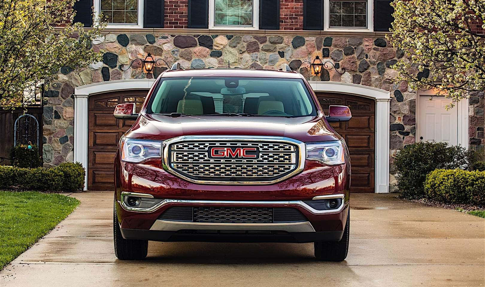 Gmc Acadia 桌布 壁紙 — 一輛光亮的棗紅色GMC Acadia SUV停放在石面房屋前的混凝土車道上，房屋有木製車庫門和綠色灌木。