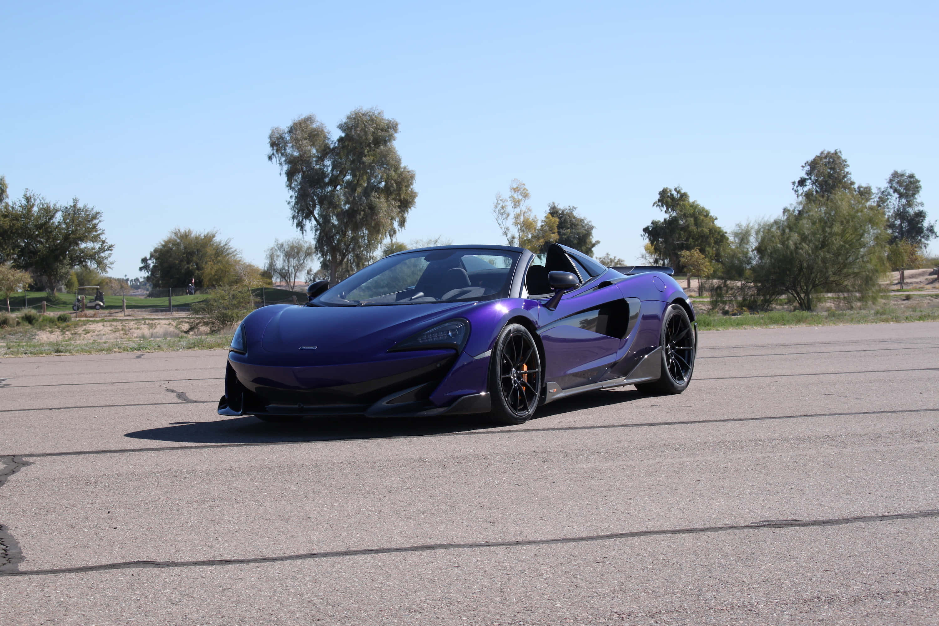 Mclaren 600lt Spider 桌布 壁紙 — 一輛光亮的紫色McLaren跑車停在瀝青路上，背景是晴朗的藍天和散落的樹木。