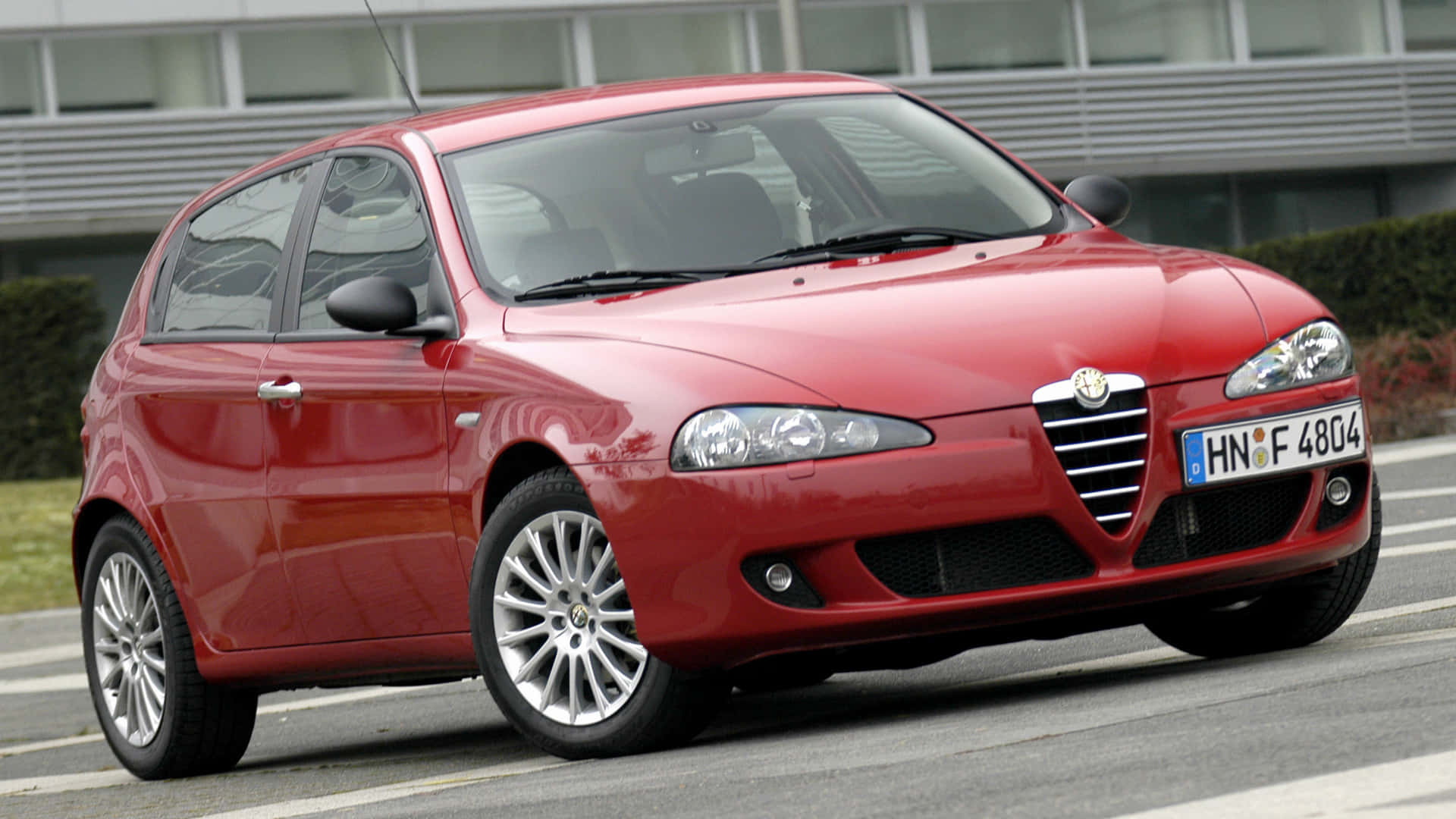 Alfa Romeo 147 桌布 壁紙 — 一輛光澤的紅色愛快羅密歐掀背車停在柏油路上，背景有現代建築和綠化。