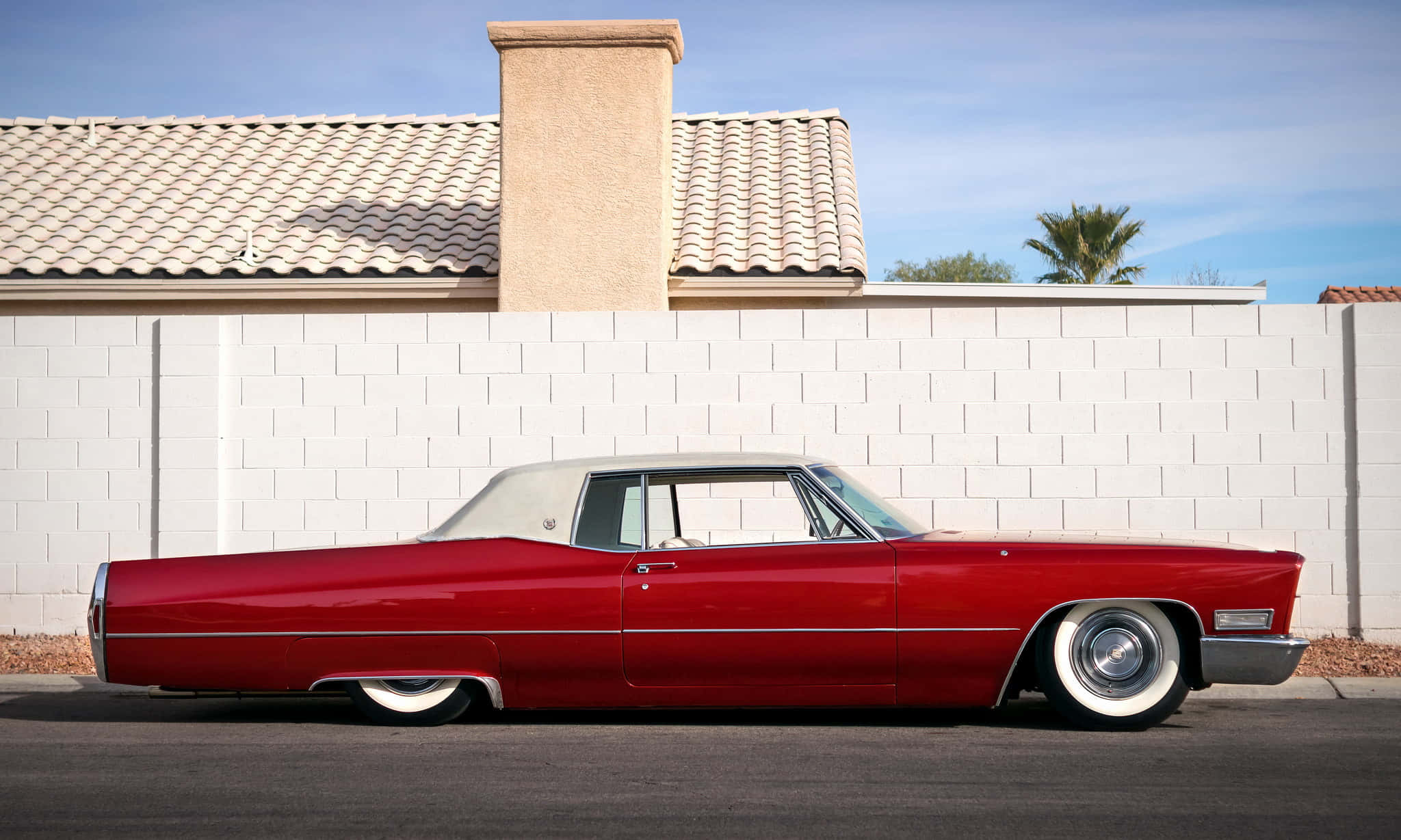 Cadillac Deville 桌布 壁紙 — 一輛光澤的紅色經典凱迪拉克，白色車頂，停在白色磚牆前，背景是晴朗的藍天。