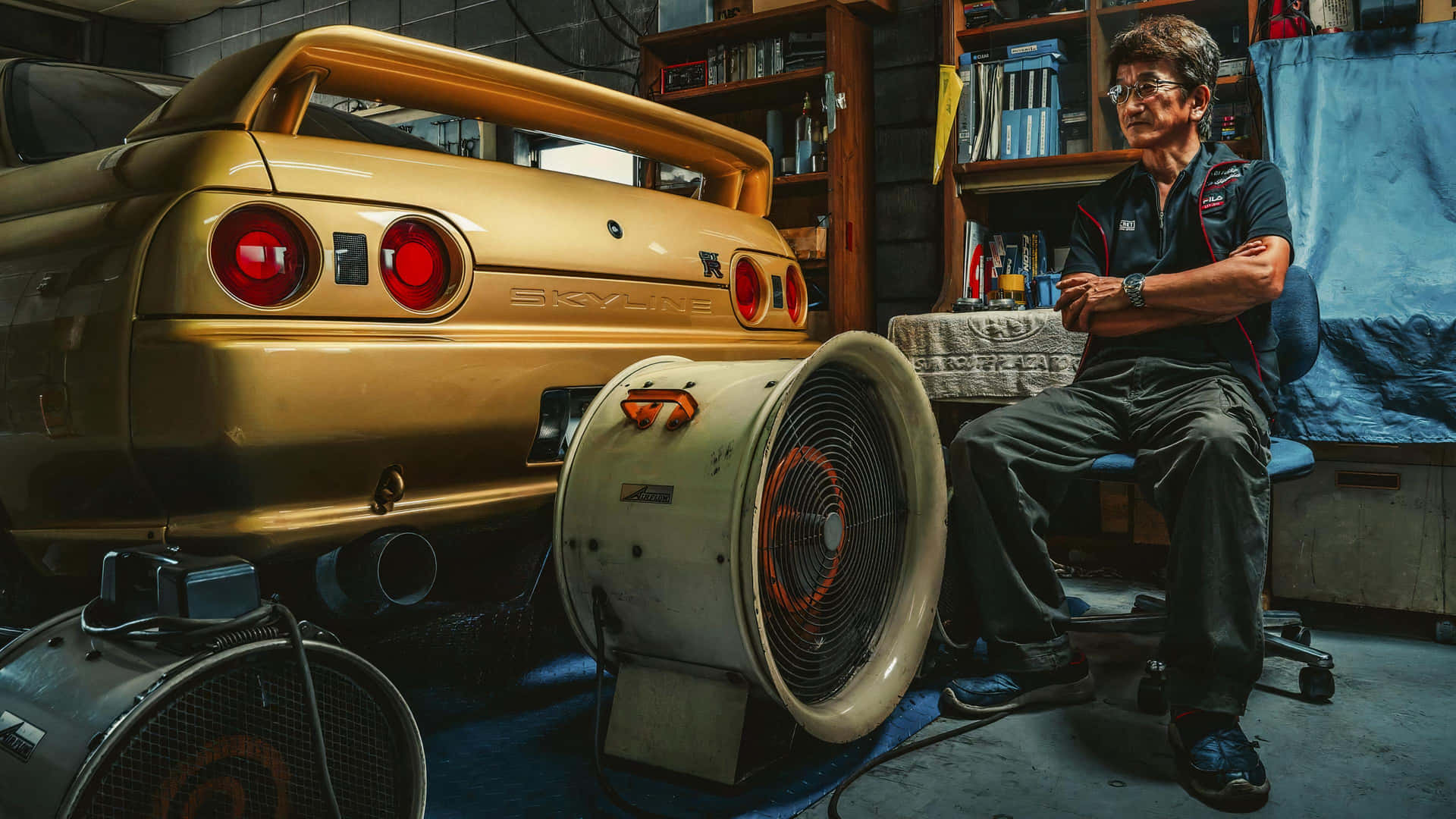  壁紙 — 一輛金色日產Skyline R34停在雜亂的車庫裡，旁邊有一個大型工業風扇，一位身穿工作服的技師坐在椅子上沉思。