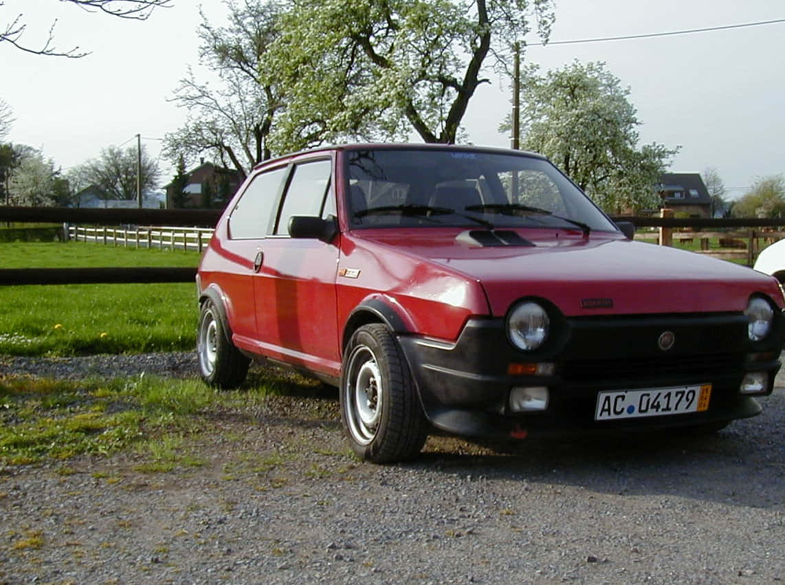 Fiat Ritmo 桌布 壁紙 — 一輛經典紅色菲亞特熊貓，帶黑色飾邊，停在碎石路上，旁邊是長滿嫩芽樹木的草地。