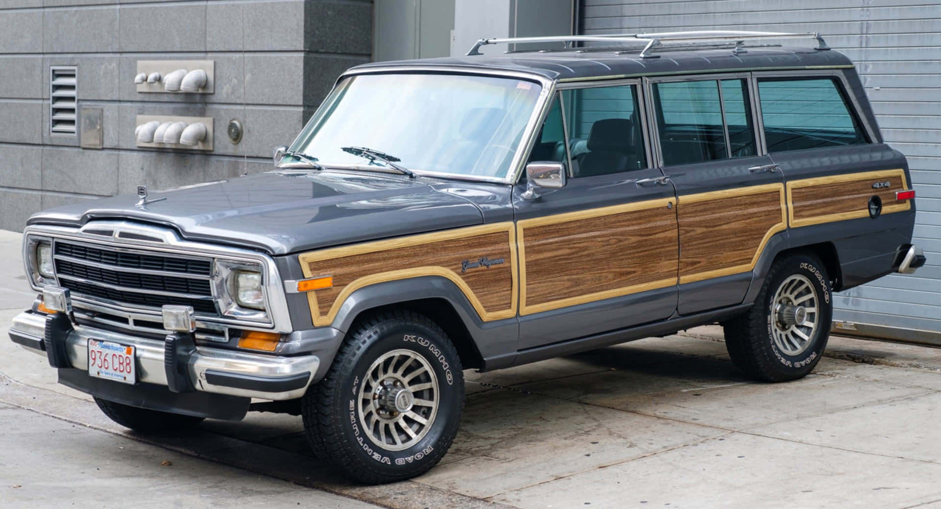 Jeep Wagoneer 桌布 壁紙 — 一輛經典灰色Jeep Wagoneer配備木紋飾板，停在灰色磚砌建築旁的混凝土人行道上。