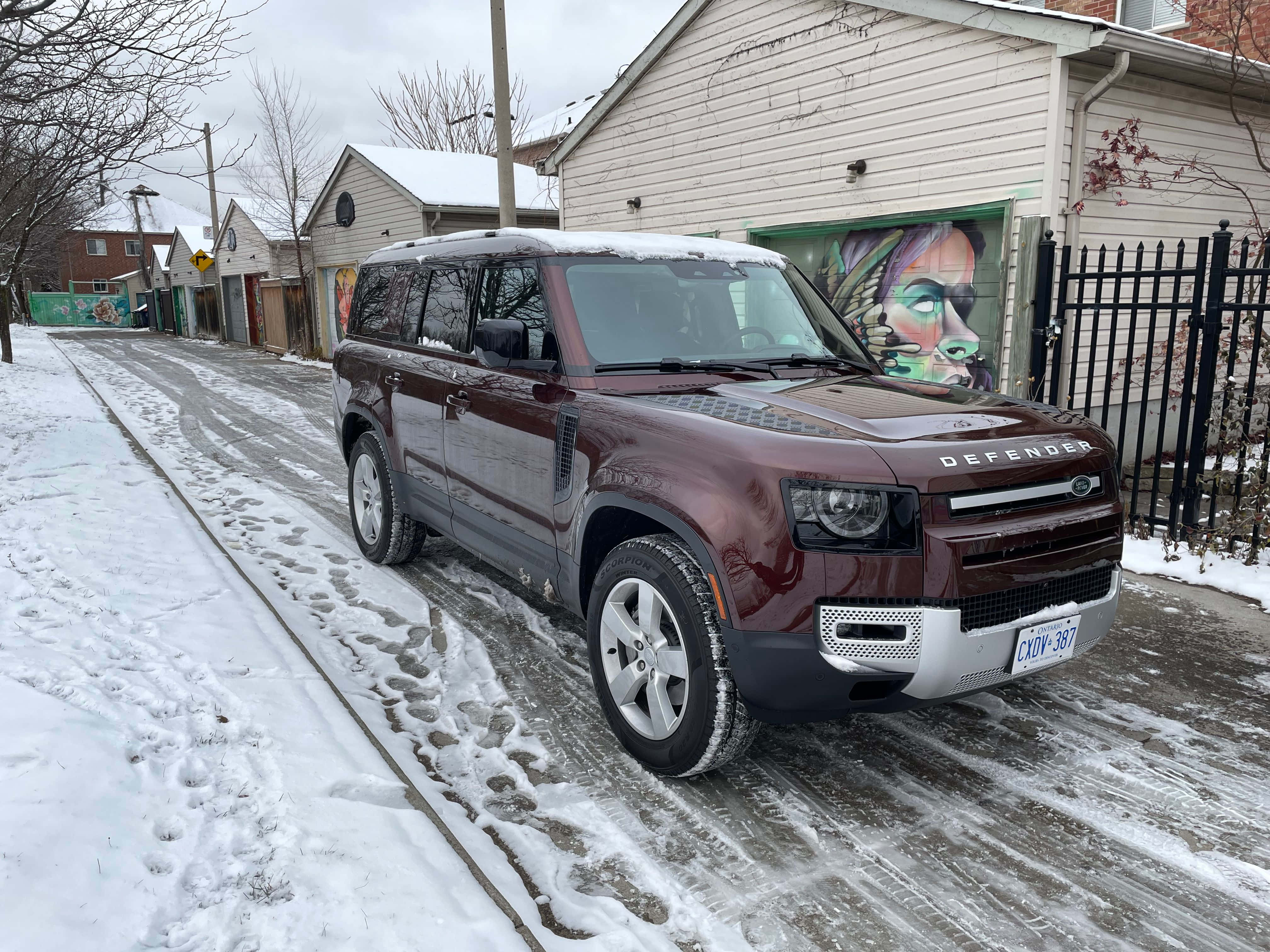 Land Rover Defender 桌布 壁紙 — 一輛酒紅色的Land Rover Defender停放在積雪的住宅街道旁，旁邊是一個佈滿塗鴉的車庫。
