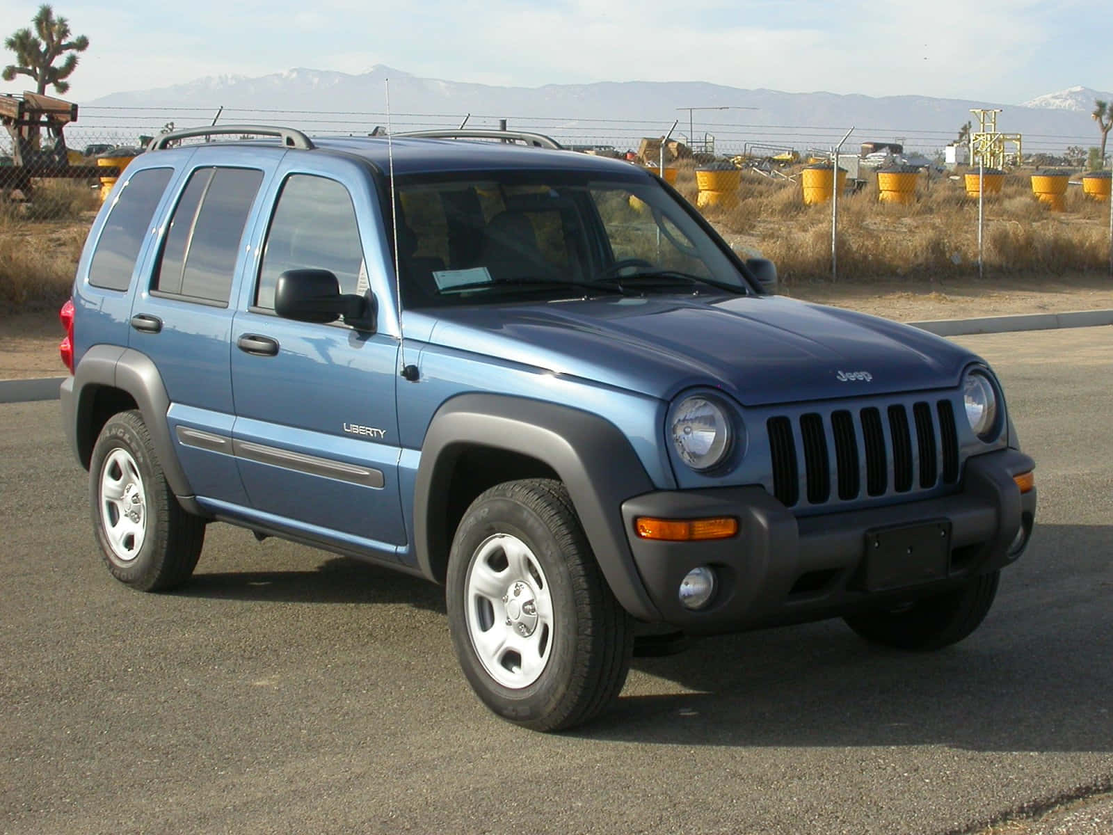 Jeep Liberty 桌布 壁紙 — 一輛藍色Jeep Liberty SUV停泊在柏油路上，背景有山脈和黃色桶，天空晴朗。