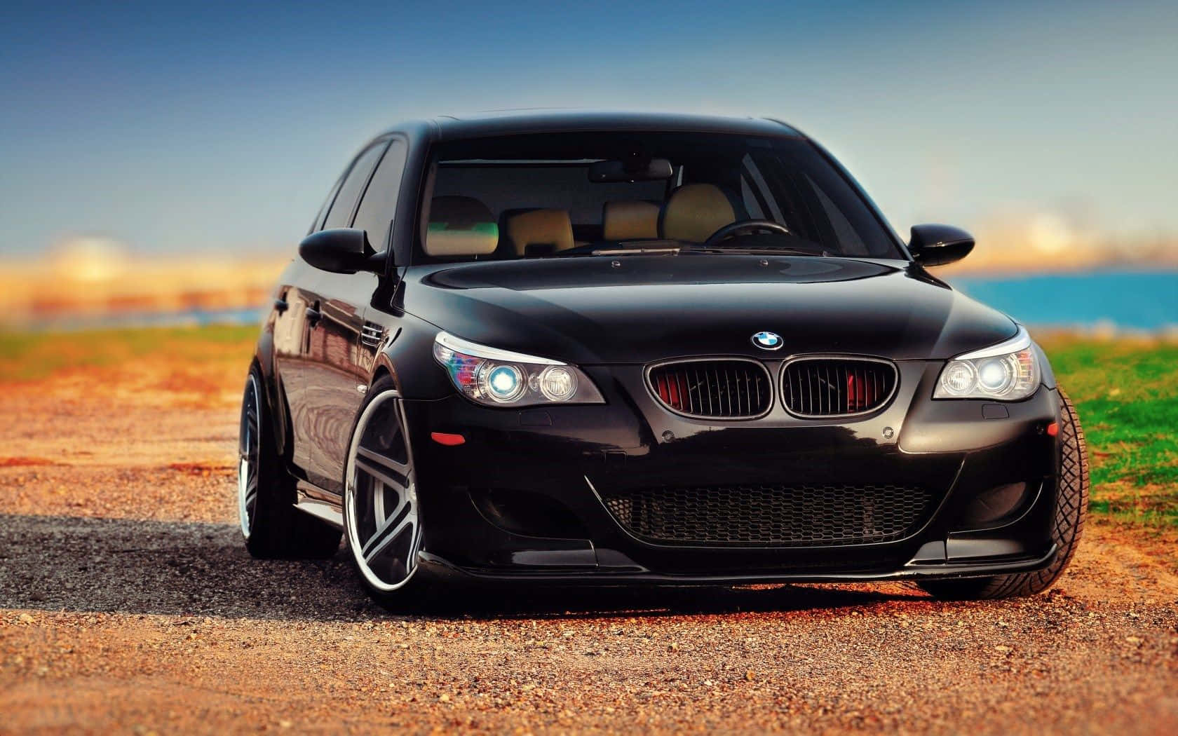 Bmw M5 桌布 壁紙 — 一輛亮黑色BMW M5運動轎車停在碎石路上，背景是模糊的海岸景觀。