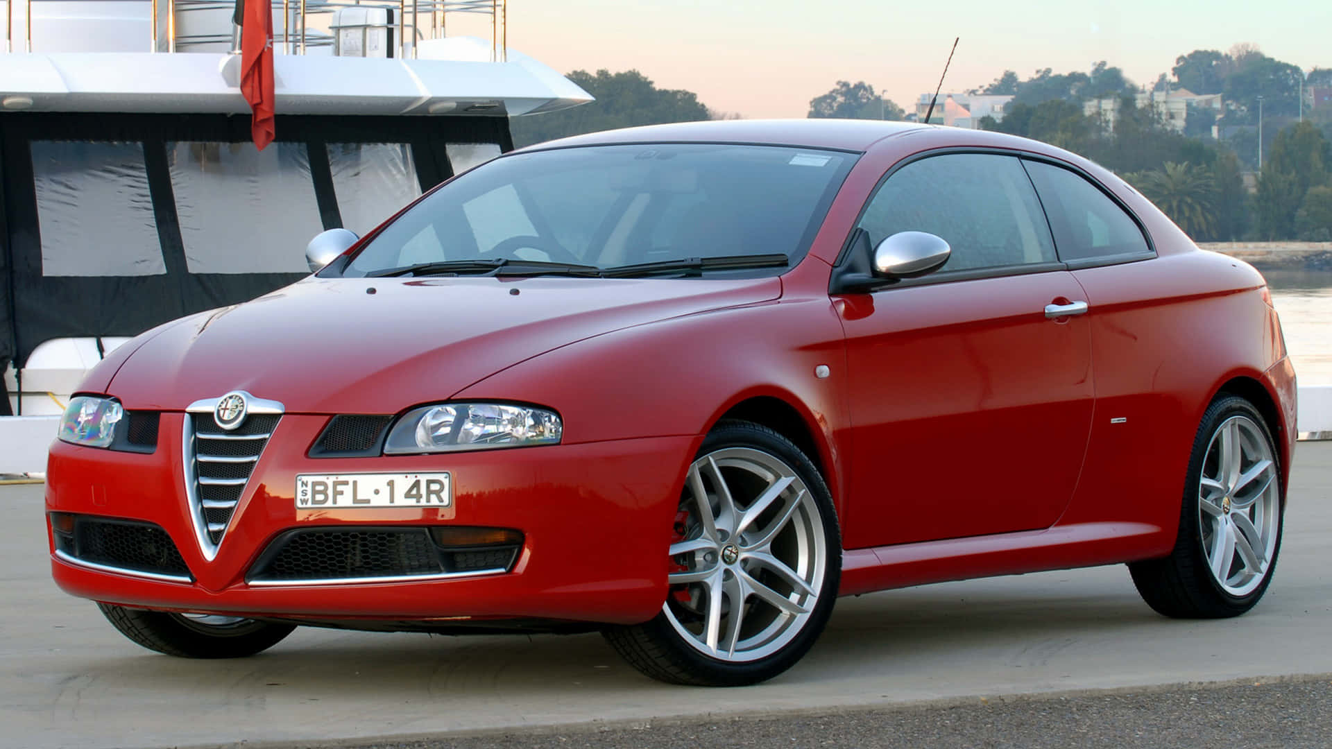 Alfa Romeo Gt 桌布 壁紙 — 一輛亮紅色愛快羅密歐轎跑車停泊在遊艇旁，夕陽西下，溫暖的環境光線反射在其流暢的曲線上。
