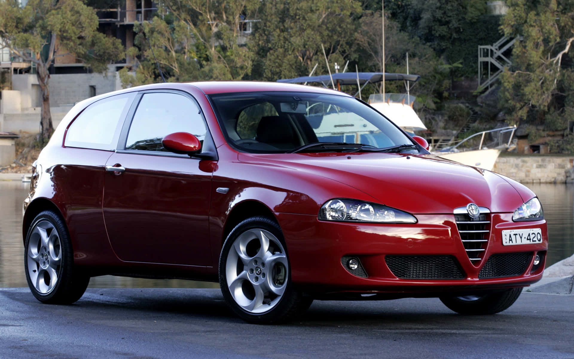 Alfa Romeo 147 桌布 壁紙 — 一輛亮紅色的愛快羅密歐147掀背車停在海濱，背景有船隻和樹木。