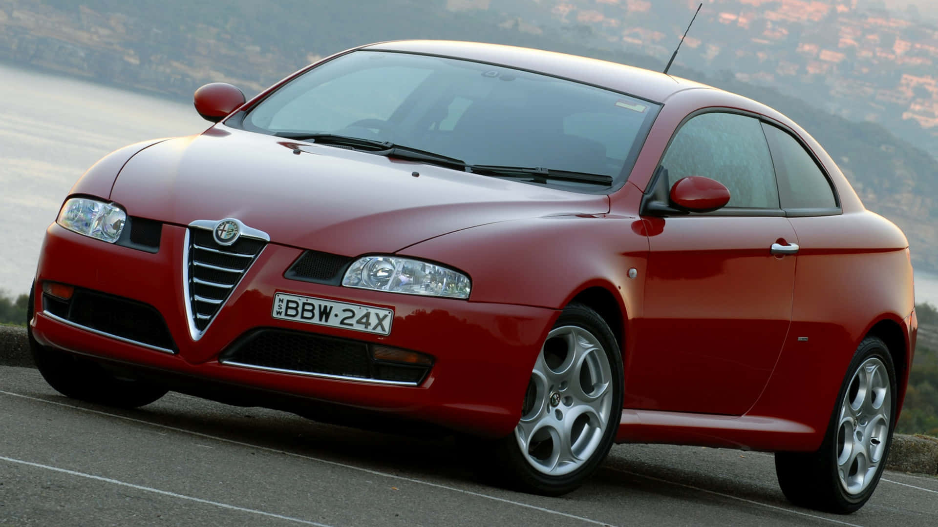 Alfa Romeo Gt 桌布 壁紙 — 一輛亮紅色的愛快羅密歐轎跑車停泊在沿海公路，背景是模糊的城市景觀。