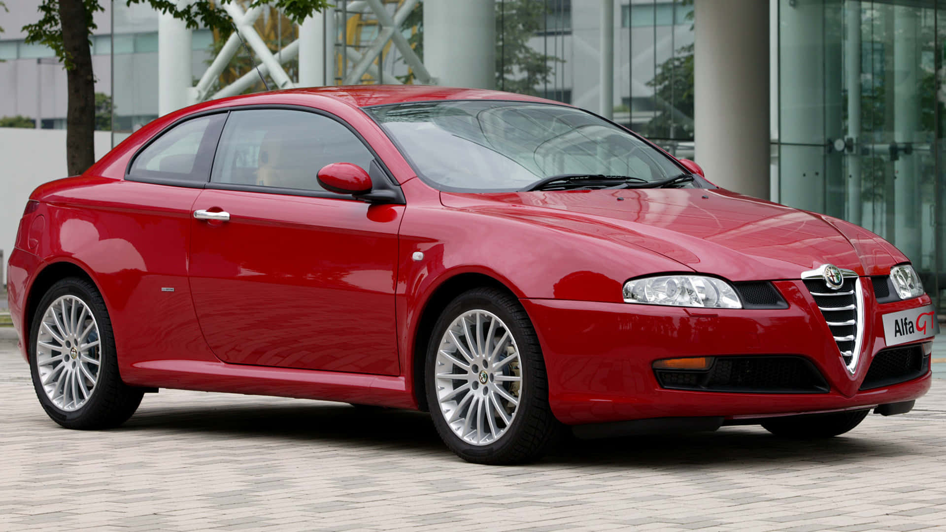 Alfa Romeo Gt 桌布 壁紙 — 一輛亮紅色的Alfa Romeo GT轎跑車停放在鋪砌的廣場上，背景是現代玻璃建築。