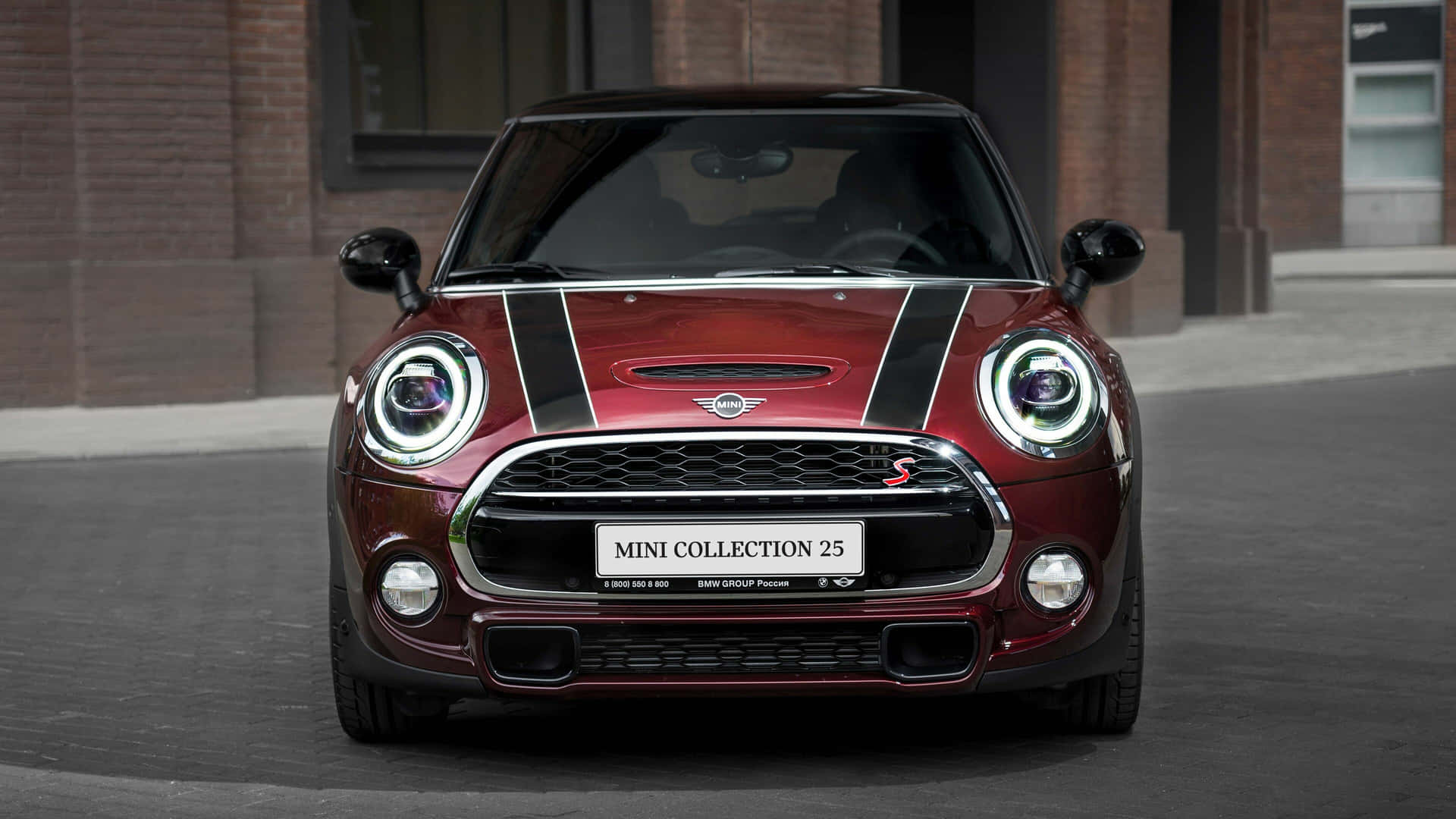  壁紙 — 一輛亮紅色MINI Cooper S帶有黑色賽車條紋，在磚砌建築前的城市街道上正面朝前。