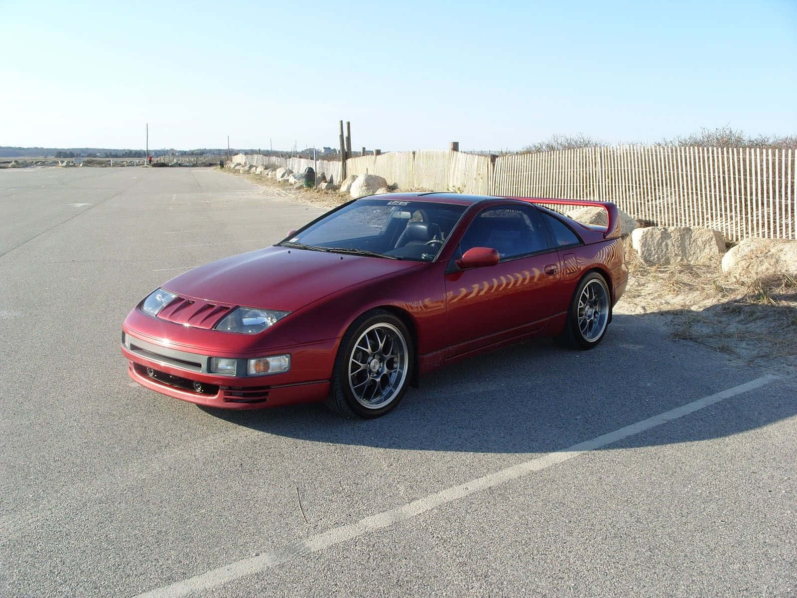 Nissan 300zx 桌布 壁紙 — 一輛亮紅色日產300ZX跑車停泊在柏油路上，旁邊有木柵欄，天空湛藍清澈。