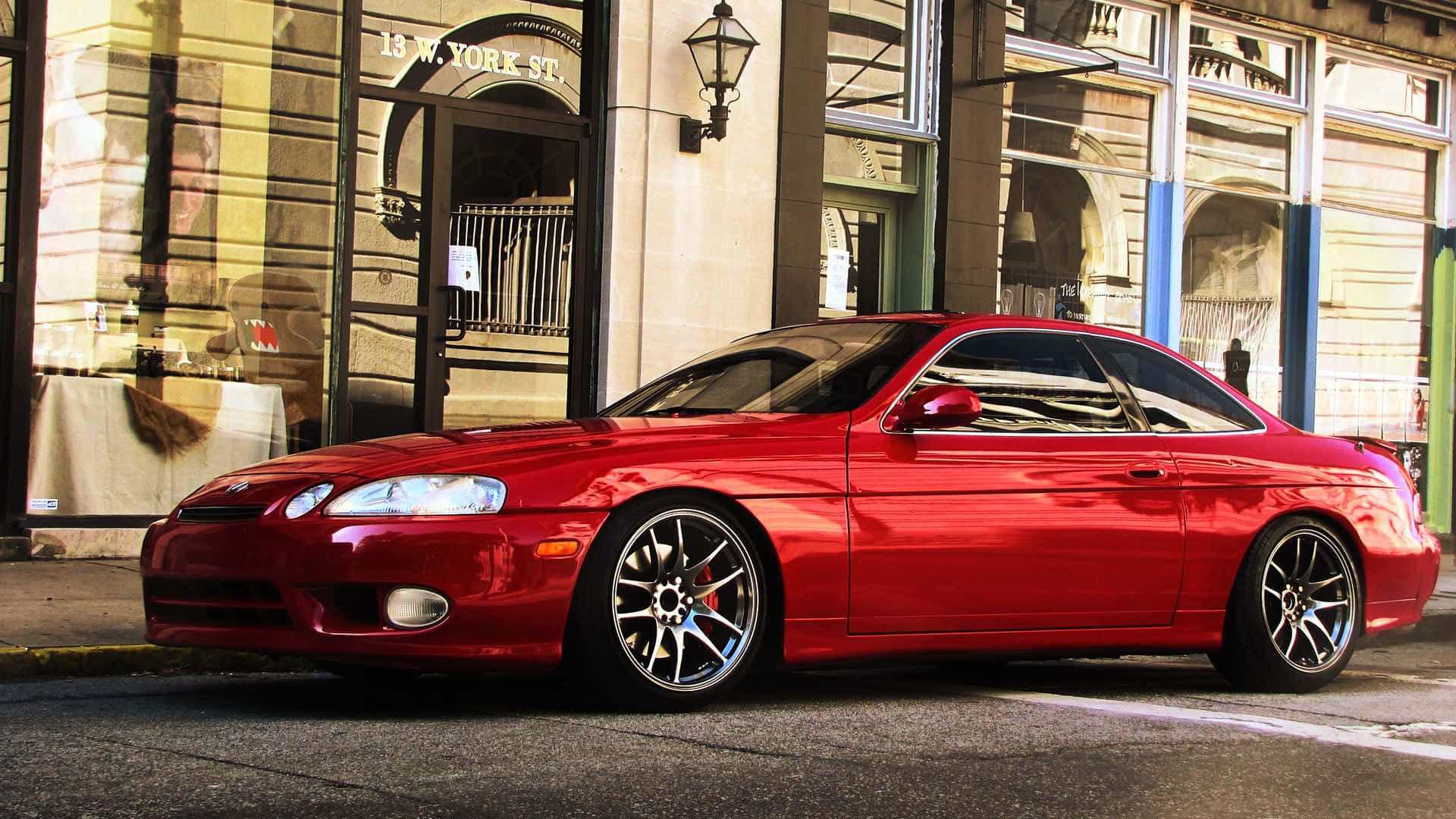 Lexus Sc 300 桌布 壁紙 — 一輛亮紅色運動型轎跑車停放在城市街道上，旁邊是擁有大型窗戶和復古路燈的歷史建築。